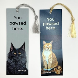 May include: Two cat-themed bookmarks with tassels. One is light gray with a black cat illustration and the text "You pawsed here." The other is navy blue with an orange tabby cat and the same text.
