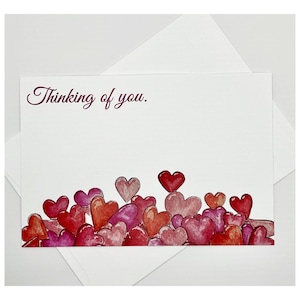 May include: A white greeting card with the text "Thinking of you." in a cursive font. The bottom of the card is decorated with watercolor hearts in shades of red, pink, and purple. An envelope is behind the card.
