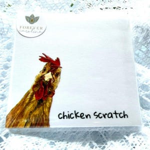 May include: A square notepad with a white background featuring a brown chicken illustration in the bottom left corner. The text "chicken scratch" is printed in black in the bottom right. A circular logo reads "FOREVER caroline" at the top left.
