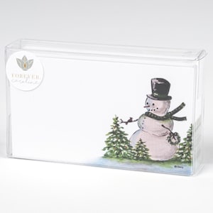 May include: A clear plastic box containing a set of cards. The cards feature a watercolor illustration of a snowman wearing a top hat and scarf, with evergreen trees in the background. The box has a sticker that says "FOREVER. Caroline."