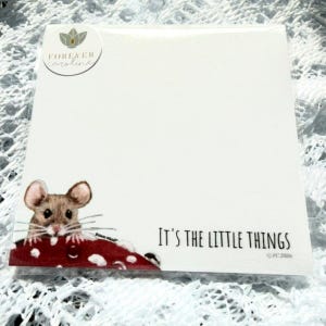 May include: A square notepad featuring a brown mouse peeking over a red and white spotted mushroom. The text "It's the little things" is printed in black on the bottom right. A circular logo for Forever Caroline is in the top left corner.