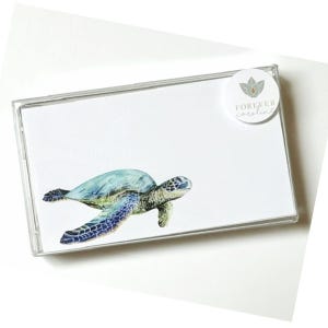 May include: A rectangular clear plastic case containing white stationery cards. Each card features a watercolor illustration of a sea turtle in shades of blue and green. A circular white sticker with a logo and the text FOREVER caroline is attached.