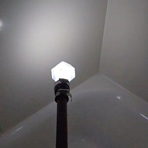 Puede incluir: Una lámpara geométrica hexagonal iluminada está montada en un poste de metal oscuro. La luz es blanca brillante y la lámpara está asegurada con una abrazadera de metal. El fondo es una pared gris claro.