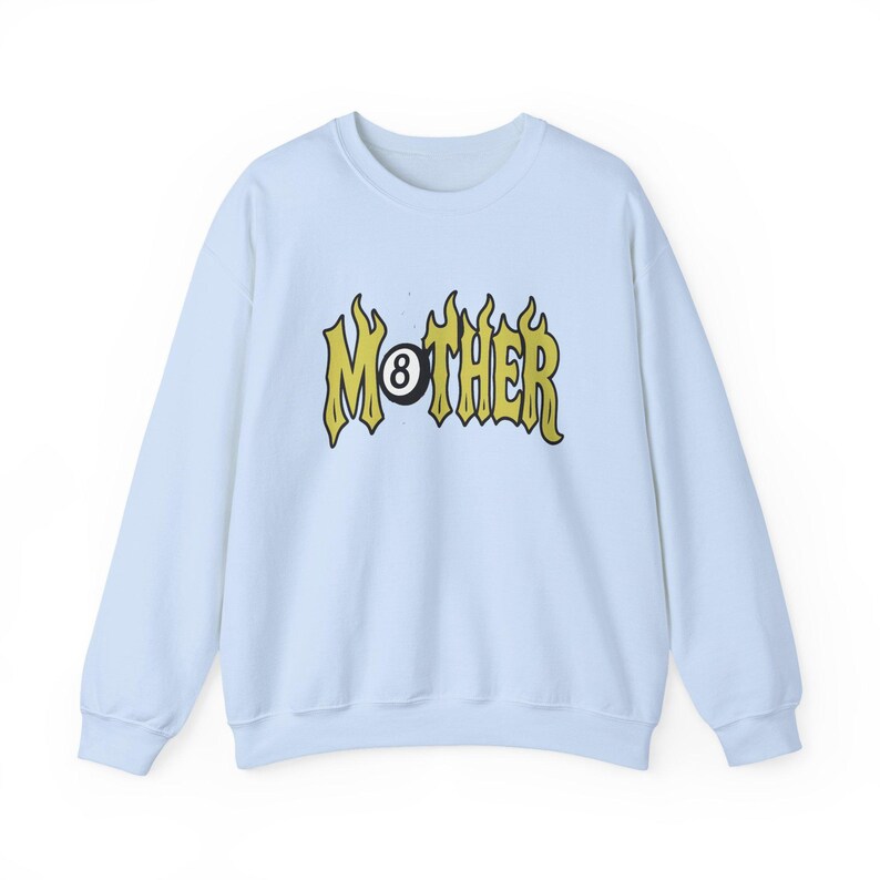 K&ouml;nnte beinhalten: Hellblaues Sweatshirt mit dem Wort "MOTHER" in gelber, flammenartiger Schrift, wobei die "O" durch eine schwarze Billardkugel ersetzt wurde. Das Sweatshirt hat lange &Auml;rmel und einen Rundhalsausschnitt.