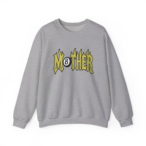 K&ouml;nnte beinhalten: Ein hellgraues Sweatshirt mit Rundhalsausschnitt. Auf der Vorderseite steht das Wort "MOTHER" in gelber, flammenartiger Schrift, wobei eine schwarze Achtkugel das "O" ersetzt. Das Sweatshirt hat lange &Auml;rmel und eine lockere Passform.