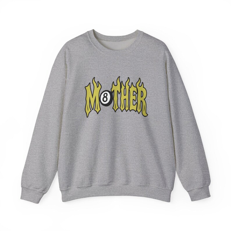 K&ouml;nnte beinhalten: Ein hellgraues Sweatshirt mit Rundhalsausschnitt. Auf der Vorderseite steht das Wort "MOTHER" in gelber, flammenartiger Schrift, wobei eine schwarze Achtkugel das "O" ersetzt. Das Sweatshirt hat lange &Auml;rmel und eine lockere Passform.