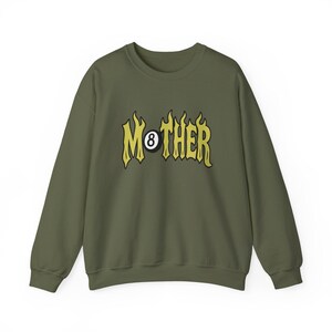 K&ouml;nnte beinhalten: Olivgr&uuml;nes Sweatshirt mit dem Wort "MOTHER" in gelben, flammenartigen Buchstaben. Der Buchstabe "O" ist durch eine 8-Ball-Grafik ersetzt. Das Sweatshirt hat einen Rundhalsausschnitt und lange &Auml;rmel.