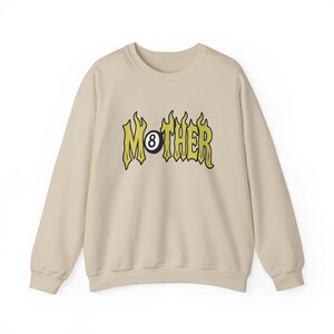 K&ouml;nnte beinhalten: Cremefarbenes Sweatshirt mit dem Wort "MOTHER" in gelber, flammenartiger Schrift, wobei eine schwarze Achtkugel das "O" ersetzt. Das lang&auml;rmlige Oberteil hat einen Rundhalsausschnitt und eine lockere Passform.