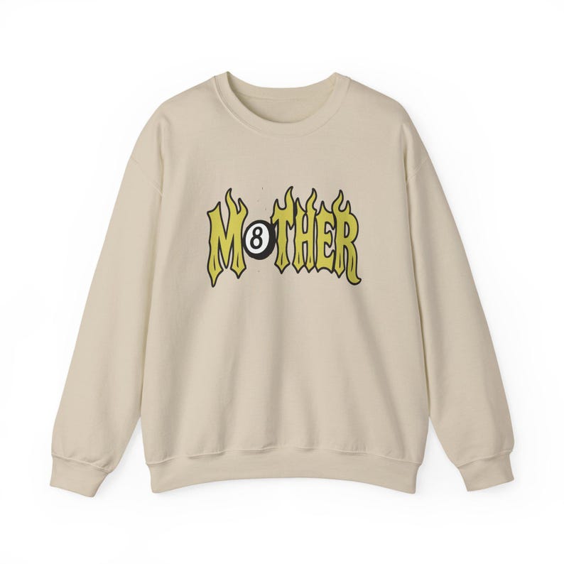 K&ouml;nnte beinhalten: Cremefarbenes Sweatshirt mit dem Wort "MOTHER" in gelber, flammenartiger Schrift, wobei eine schwarze Achtkugel das "O" ersetzt. Das lang&auml;rmlige Oberteil hat einen Rundhalsausschnitt und eine lockere Passform.
