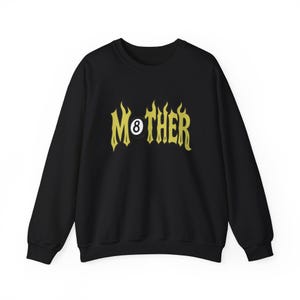 K&ouml;nnte beinhalten: Schwarzes Sweatshirt mit dem Wort "MOTHER" in gelber, flammenartiger Schrift. Die Zahl 8 ist in das Design integriert. Das Sweatshirt hat einen Rundhalsausschnitt und lange &Auml;rmel.