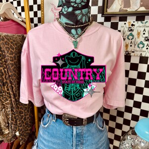 Puede incluir: Camiseta rosa con un gr&aacute;fico de ne&oacute;n que dice "COUNTRY STATE OF MIND". La camiseta se combina con pantalones cortos de mezclilla, un cintur&oacute;n negro con hebilla plateada y una bandana turquesa y negra.