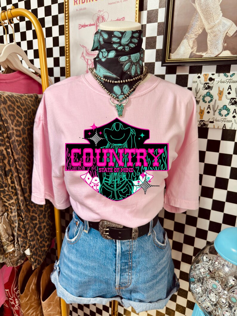 Puede incluir: Camiseta rosa con un gr&aacute;fico de ne&oacute;n que dice "COUNTRY STATE OF MIND". La camiseta se combina con pantalones cortos de mezclilla, un cintur&oacute;n negro con hebilla plateada y una bandana turquesa y negra.