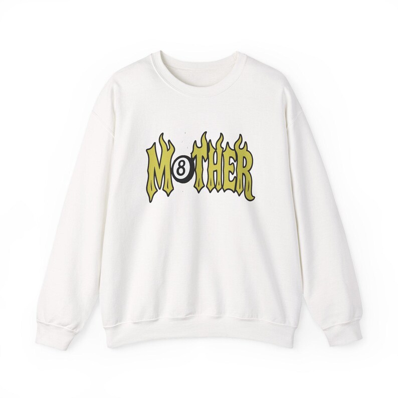 K&ouml;nnte beinhalten: Wei&szlig;es Sweatshirt mit Rundhalsausschnitt und dem Wort "MOTHER" in einer flammenartigen Schriftart, mit einer Acht-Ball-Kugel in der Mitte. Der Text ist gelb und schwarz. Das Sweatshirt ist ein l&auml;ssiger Stil.