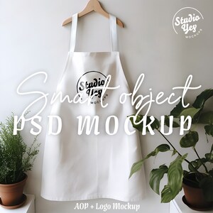May include: White apron with a black logo that says "Studio Yes Mockups" on the front. The apron is hanging on a wooden hanger against a white wall. There are two potted plants on either side of the apron.