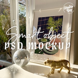 Op de afbeelding: Een wit ingelijst beeld met een kleurrijke abstracte print van een plant en fruit. Een gouden tijgerfiguur staat op een wit oppervlak naast een zilveren discobal.