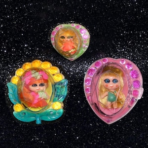 May include: Three vintage doll pendants. Each doll is encased in a clear plastic bubble. One is heart-shaped, one is flower-shaped, and the other is heart-shaped. The dolls have blonde or orange hair and wear colorful outfits.