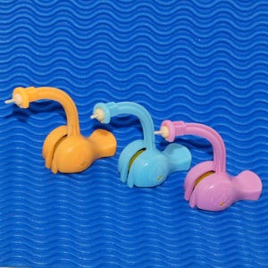 May include: Three colorful, plastic baby bottle holders in orange, blue, and pink. Each has a curved neck and a base with a small, circular design. The background is a textured, blue surface.