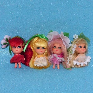May include: Four small dolls with distinct hair colors and floral-themed outfits. The first has red hair and a red dress, the second has yellow hair and a white lace dress, the third has pink hair and a pink lace dress, and the fourth has blonde hair and a white dress.