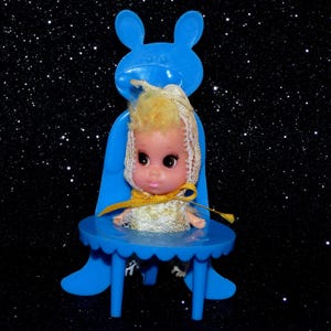 May include: A vintage doll with blonde hair and large eyes, wearing a white and gold dress and a lace head covering. The doll is seated in a blue plastic chair with a bunny ear design on the back. The chair has a round table.