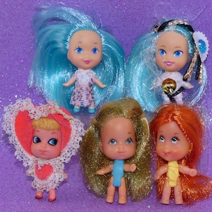 May include: Five small dolls with distinct hair colors and outfits. Two dolls have bright blue hair, one wears a pink patterned dress, another a white outfit with a black belt. One doll wears a red heart-shaped bonnet, one has blonde hair, and one has orange hair.