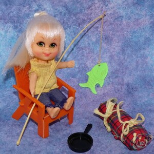 May include: A vintage doll with white hair, wearing a yellow top and blue jeans, sits in an orange chair, fishing with a green fish-shaped lure. A rolled-up red plaid blanket, a black pan, and a fishing rod complete the scene.