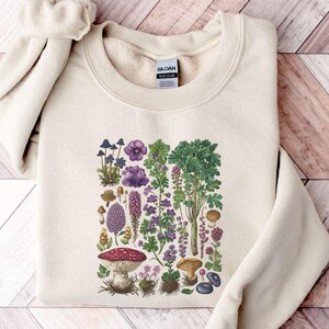 May include: Cream-colored sweatshirt featuring a botanical illustration of various mushrooms and plants in shades of purple, green, and brown. The design includes a large red and white mushroom, along with other fungi and floral elements. The sweatshirt has a crew neck and long sleeves.