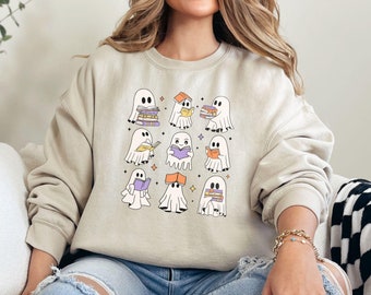 Puede incluir: Una sudadera beige de cuello redondo con un estampado gráfico de nueve fantasmas blancos, cada uno interactuando con libros coloridos. Los fantasmas aparecen en varias posturas, algunos sosteniendo pilas de libros y otros leyendo, con estrellas.
