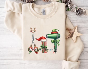 Op de afbeelding: Crèmekleurige sweatshirt met een kerstthema-ontwerp voor een kapper. Het ontwerp omvat een schaar met rendiergewei, een kam met een kerstmuts en een groene föhn met een elfenmuts. Het ontwerp is versierd met kerstverlichting en strikken.