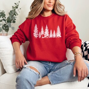 May include: A red sweatshirt with a white silhouette of a row of pine trees. The sweatshirt is worn by a person sitting on a white couch.