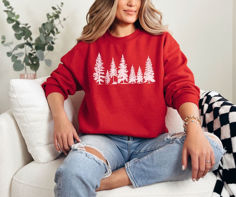 May include: A red sweatshirt with a white silhouette of a row of pine trees. The sweatshirt is worn by a person sitting on a white couch.