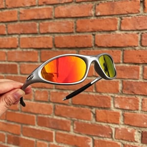 May include: Silver-toned sunglasses featuring a wrap-around frame design and reflective lenses that shift from orange to yellow. The temples are black, and the brand name Oakley is printed in small black letters on the upper corner of the lens.