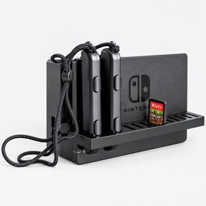 May include: A black Nintendo Switch dock featuring a horizontal shelf holding two black Joy-Con controllers with attached black wrist straps and a single Minecraft game cartridge. The dock displays the Nintendo Switch logo on its front panel.