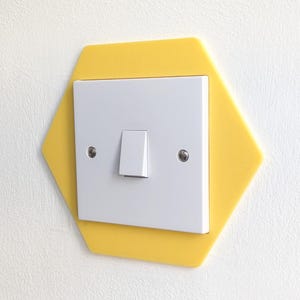 May include: A white light switch with a single toggle, set within a bright yellow, hexagonal surround. The surround is mounted on a white wall. The switch has two small silver screws.