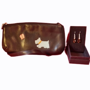 May include: A dark brown leather clutch bag with a zip and a butterfly detail. A white dog with a yellow collar is also on the bag. A pair of teardrop earrings in a dark box are next to the bag.