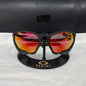 May include: Matte black sunglasses featuring a wrap-around frame design and reflective red-orange lenses. The eyewear is mounted on a black display stand with the gold-coloured Oakley logo printed on the base, showcasing a sleek, athletic aesthetic.