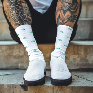 May include: White socks with a pattern of small teal fish. The socks are worn with white slip-on shoes with black soles. The image shows the lower legs and feet of a person sitting on concrete steps.