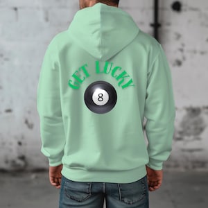 Puede incluir: Una sudadera con capucha verde claro con las palabras "GET LUCKY" en verde arqueadas sobre un gráfico de bola 8 en blanco y negro. La sudadera es de color sólido y tiene una capucha.
