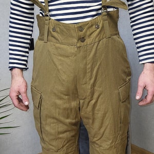 Op de afbeelding: Een bruine cargo broek met bretels. De broek heeft een hoge taille en is gemaakt van een dikke, duurzame stof. De bretels zijn verstelbaar en hebben een metalen gesp.