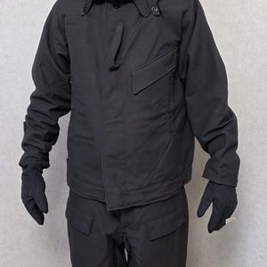 Puede incluir: Conjunto de chaqueta y pantalón negros con cubrecara. La chaqueta tiene cierre de botón, bolsillo en el pecho y capucha. El pantalón tiene bolsillos. El cubrecara tiene agujeros para los ojos. El conjunto es todo negro.