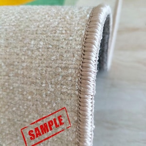 May include: Close-up of a beige carpet with a textured surface and a stitched border. The word "SAMPLE" is stamped in red on the carpet. The carpet has a colorful design in the background.