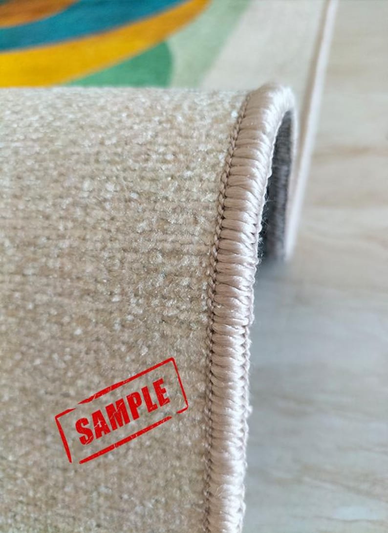 May include: Close-up of a beige carpet with a textured surface and a stitched border. The word "SAMPLE" is stamped in red on the carpet. The carpet has a colorful design in the background.
