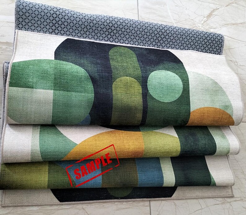 May include: Folded rugs with geometric designs in shades of green, blue, and gold. The top rug has a gray diamond pattern. The rugs are stacked, showcasing the colorful patterns and textures. The word "SAMPLE" is stamped on one of the rugs.