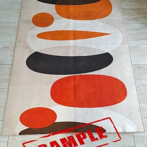 May include: A rectangular area rug featuring a beige background with a stacked pattern of overlapping circles and ovals in shades of orange, dark brown, and white. The word SAMPLE is printed in red capital letters across the bottom section.