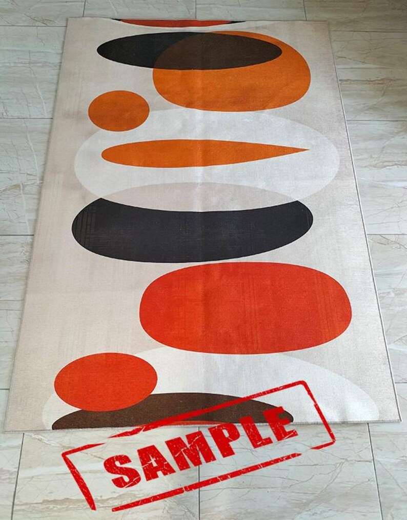 May include: A rectangular area rug featuring a beige background with a stacked pattern of overlapping circles and ovals in shades of orange, dark brown, and white. The word SAMPLE is printed in red capital letters across the bottom section.