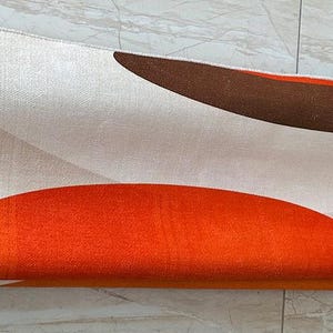 May include: A rolled area rug featuring a cream-colored background with large, abstract geometric shapes in burnt orange and dark brown. The rug displays a printed red rectangular stamp in the upper left corner containing the word SAMPLE.