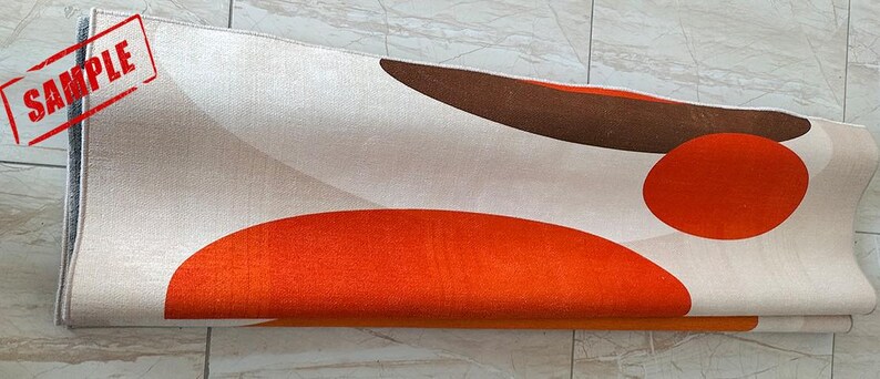 May include: A rolled area rug featuring a cream-colored background with large, abstract geometric shapes in burnt orange and dark brown. The rug displays a printed red rectangular stamp in the upper left corner containing the word SAMPLE.