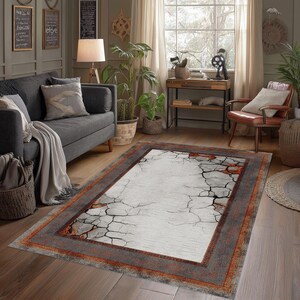 May include: A rectangular area rug featuring a central white field with a black cracked earth pattern and orange accents, surrounded by a wide, textured border in shades of dark gray and burnt orange, laid flat on a wooden floor in a living room.