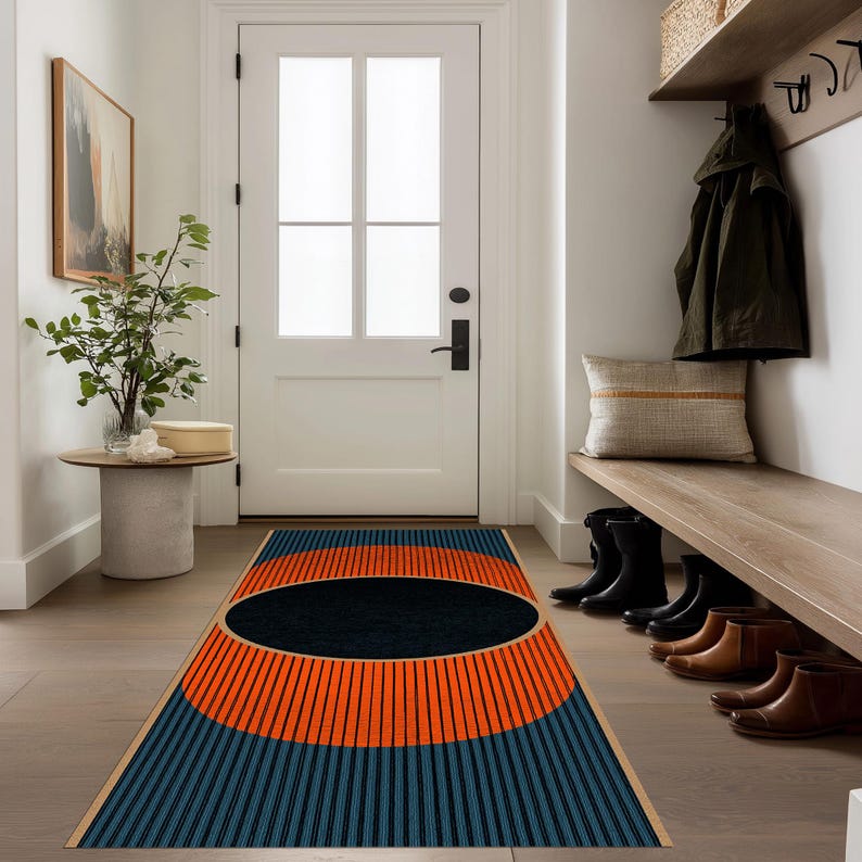 May include: A rectangular entryway rug with a geometric design. The rug features a large black oval in the center, surrounded by orange and blue sections with vertical stripes. The rug is placed in front of a white door.