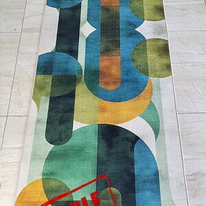 May include: A rectangular rug with an abstract design featuring overlapping shapes in shades of green, blue, orange, and yellow on a white background. The rug has a geometric pattern and a "SAMPLE" stamp in red.