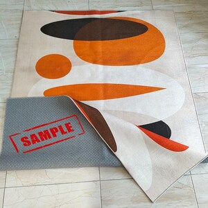 May include: A rectangular area rug featuring an abstract pattern of overlapping orange, brown, and white circular and oval shapes. The rug is partially folded to reveal a gray, textured, non-slip backing with the red text SAMPLE printed on it.